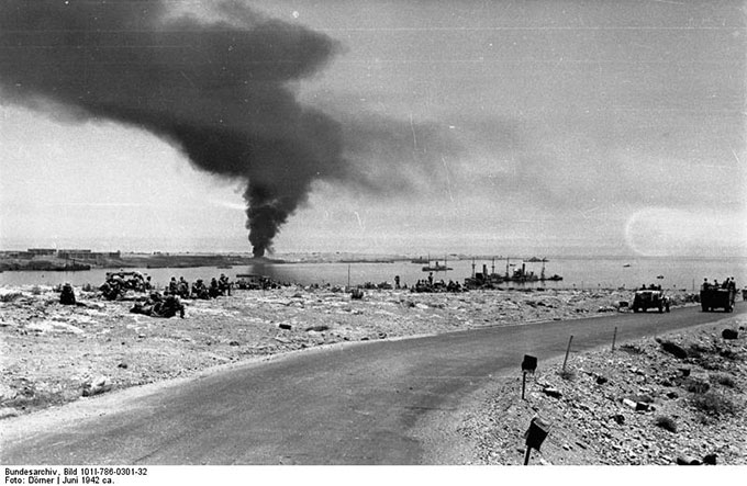 The German capture of Tobruk, with British prisoners on the left, sunken ships in the harbor and smoke over the port.